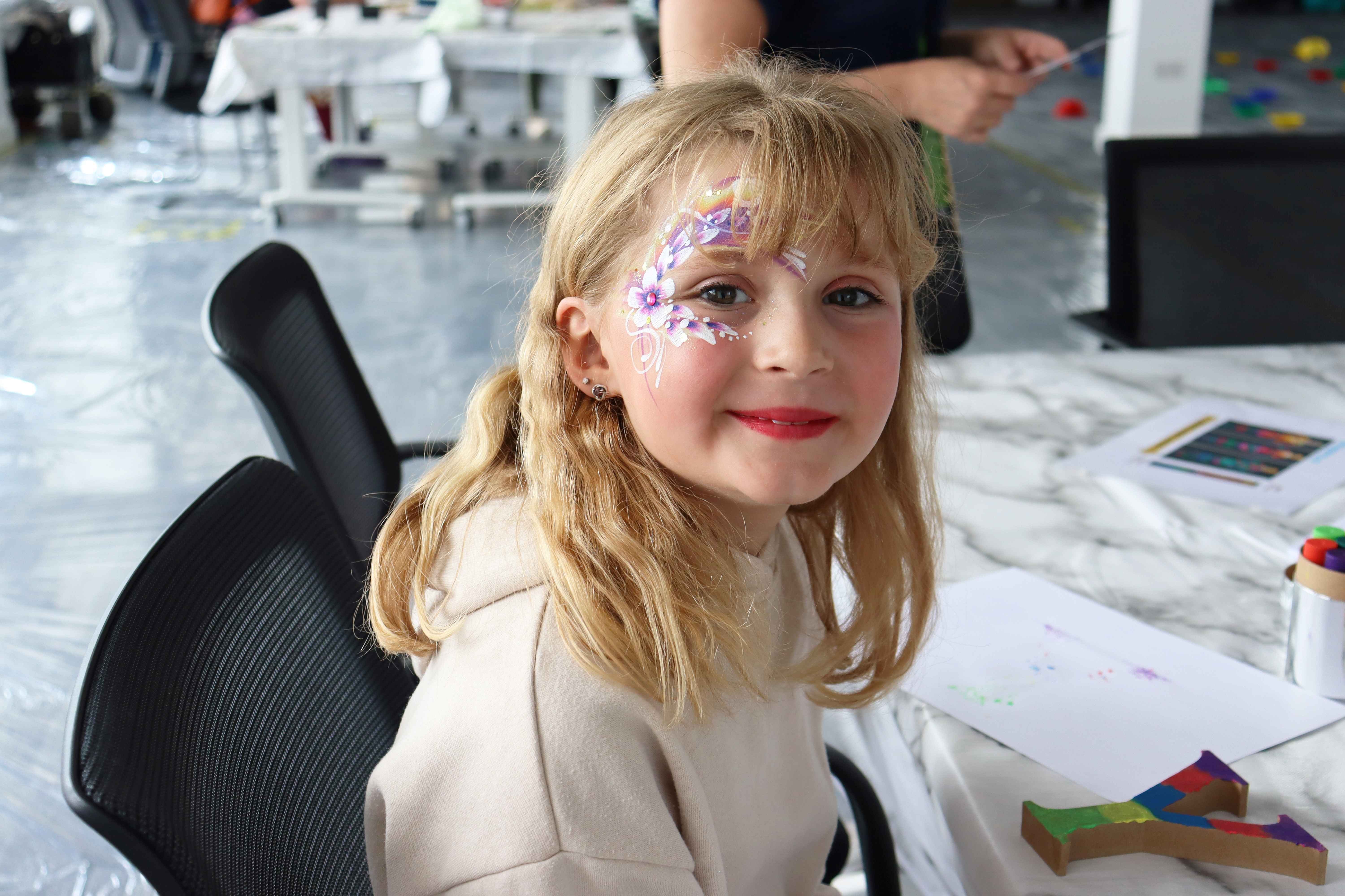 A young girl with blonde hair smiling at the camera, with a flower face paint wrapped around her left eye