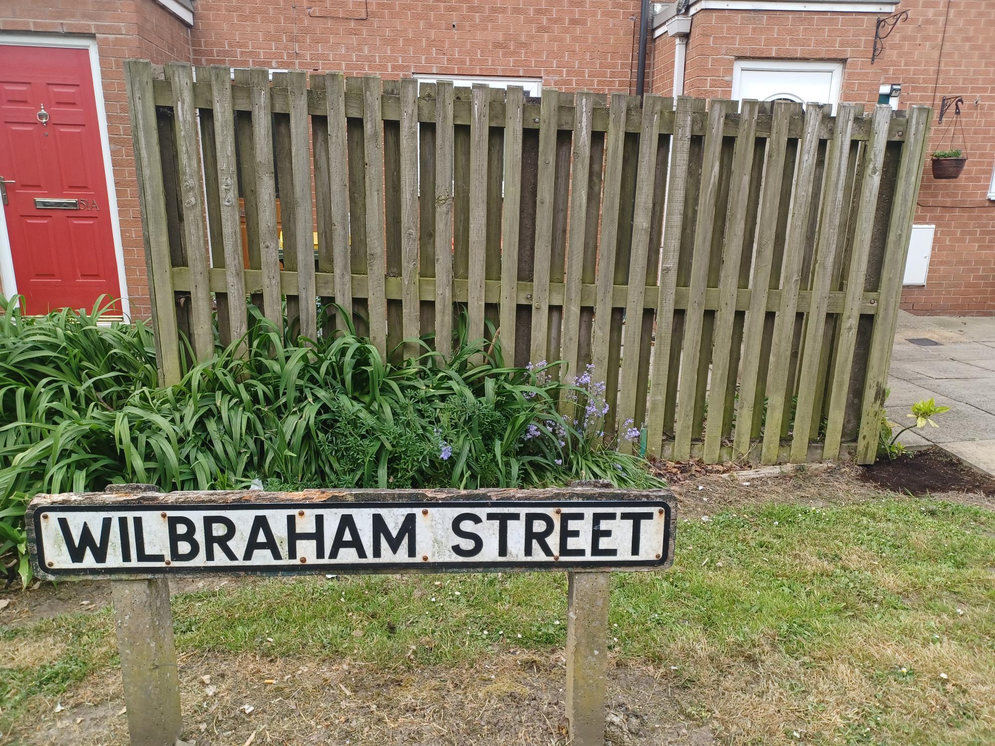 A before image of a fence with a street sign in front saying Wilbraham street