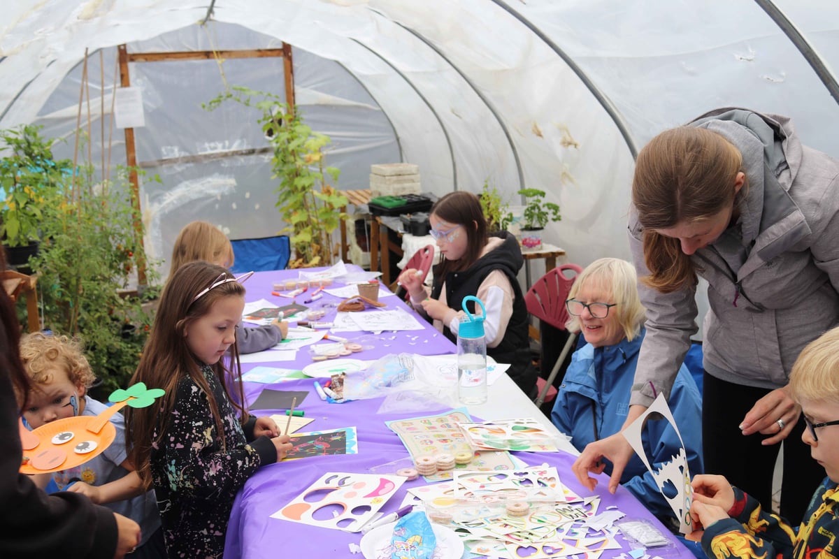 A group of adults and children sat around a purple table inside a polytunnel doing arts and crafts
